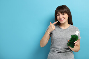 Young woman with mouthwash pointing at healthy teeth on light blue background, space for text