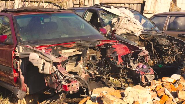 Close-up view of a damaged, abandoned car with missing parts, rusted and decrepit, among other vehicle remains in a junkyard.