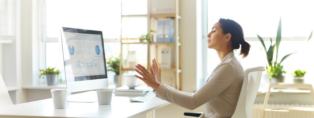 A woman is sitting at a desk with a computer monitor in front of her. She is wearing a white shirt and she is focused on her work. The room is decorated with potted plants and a vase, giving it a calm