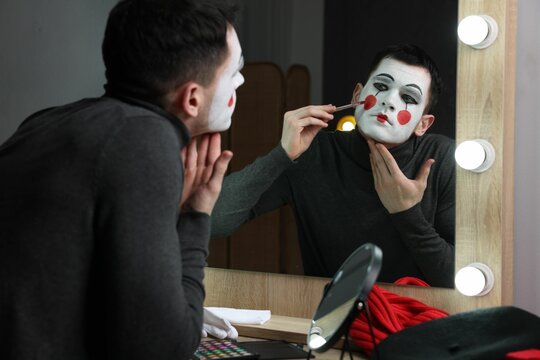 Young man applying mime makeup near mirror in dressing room