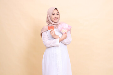 Asian Muslim woman in hijab smiling cheerfully holding rupiah cash and credit cards in crossed hands. Lifestyle, religious, business and finance concepts