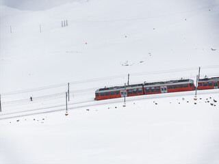 Aerial shot a bright red train glides through snowy Zermatt, beside a lone figure walking near the tracks. Sparse ski signs and red flags dot the serene landscape.