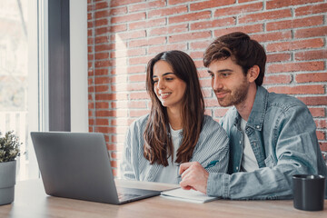 Happy millennial couple sit at table at home browsing web on laptop shopping online together,...