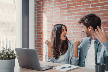Overjoyed millennial couple sit at desk look at laptop screen feel euphoric with online win,...