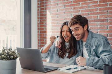 Overjoyed millennial couple sit at desk look at laptop screen feel euphoric with online win,...
