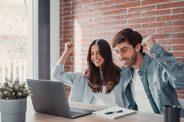 Overjoyed millennial couple sit at desk look at laptop screen feel euphoric with online win,...