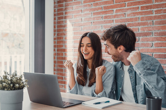 Overjoyed millennial couple sit at desk look at laptop screen feel euphoric with online win, excited happy young husband and wife read good news, get unexpected notification on computer, luck concept.