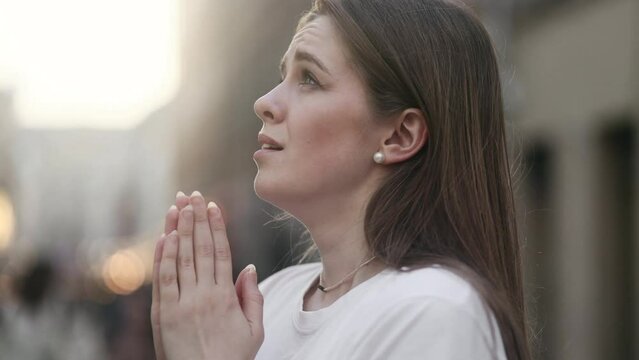 Portrait of hopeful young woman clasping hands in prayer asking for blessing and help while the rays of rising sun fall on her face at city outdoors Religion and faith concept