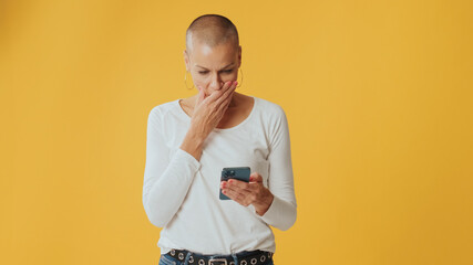 Surprised hairless woman,with phone in hands, wow effect, isolated on yellow background in studio