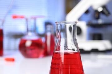 Laboratory analysis. Glass flask with red liquid on white table indoors, closeup