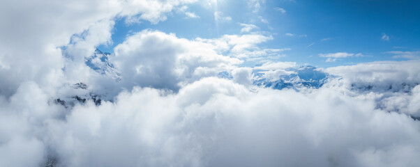 An aerial view over Verbier, Switzerland, reveals dense clouds and mountain peaks. The vivid blue...