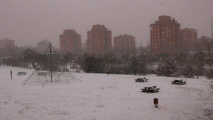 snowing in park with rope pyramid and wooden tables and brick buildings - 774519055