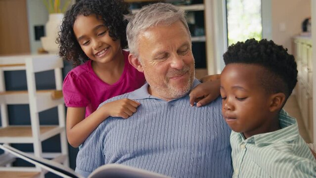 Grandfather in multi-racial family sitting with grandchildren on sofa at home reading book together - shot in slow motion