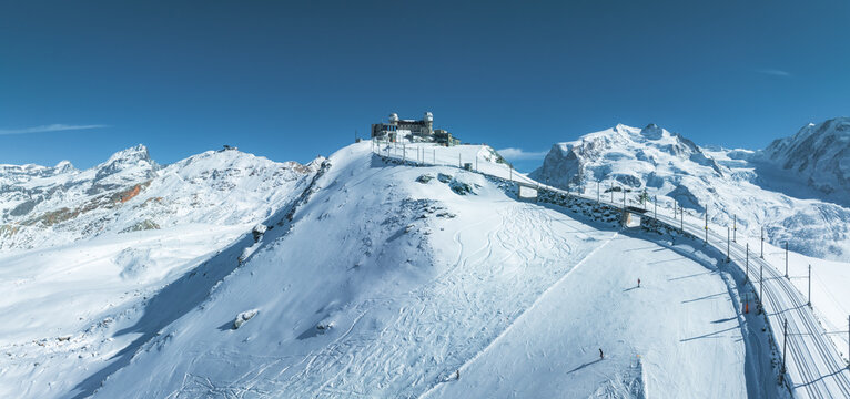 An aerial view of Zermatt ski resort in Switzerland reveals a train climbing snowy slopes, a peak facility, and skiers on thick snow. Clear skies and alpine peaks offer a stunning backdrop.