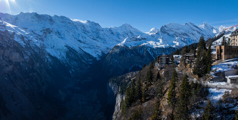 Aerial view of Murren, Switzerland, showcases a serene mountain village with traditional chalet style buildings on a cliff. Snow covered Swiss Alps and clear skies create a picturesque backdrop.