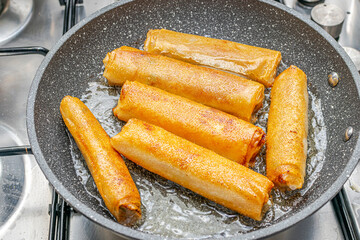 Whole pieces of Algerian boureks fried in oil and gilded in a mottled black frying pan on a hot plate on a domestic stovetop. Directly above view close-up details with reflective lights.