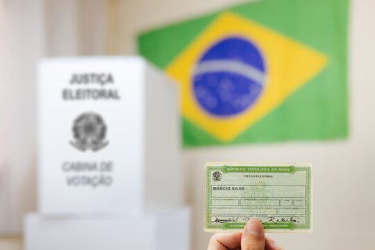 Mão com o título de eleitor, essencial para votar nas eleições do Brasil. No fundo, há uma cabine de votação da urna eletrônica e a bandeira do Brasil. Eleições e a democracria brasileira.