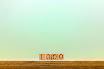 薄い青緑の背景で机の上に置かれた1000の数字のブロックの正面 - obrazy, fototapety, plakaty