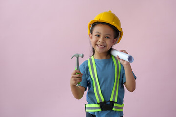 A cute girl wearing a yellow construction hat or safety helmet standing on a pink background holding a blueprint. Construction equipment, design, future career concepts, engineers, architects.