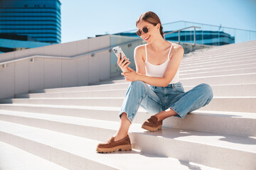Fototapeta na wymiar Young beautiful smiling hipster woman in trendy summer casual clothes. Carefree woman posing in the street. Positive model holds mobile phone, looks at cellphone screen, uses smartphone apps