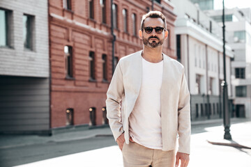 Handsome confident stylish hipster lambersexual model. Sexy modern man dressed in elegant white suit jacket. Fashion male posing in the street background in Europe city at sunset. In sunglasses - obrazy, fototapety, plakaty