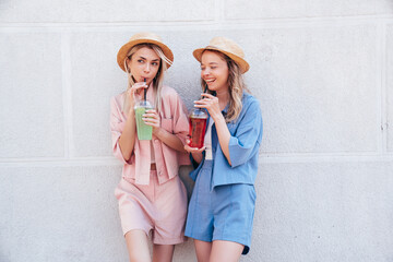 Two young beautiful smiling hipster female in trendy summer clothes. Carefree women posing outdoors. Positive models holding and drinking fresh cocktail smoothie drink in plastic cup with straw