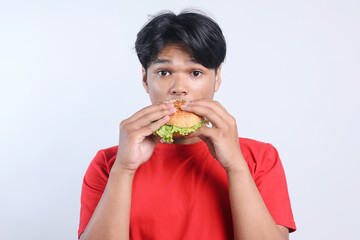 Young Asian man eating burger and looking at camera with surprise expression