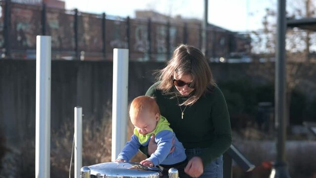 In the park, Mother and one year old red hair boy play the outdoor musical instrument Babel Drums, laughing happily, musical joy, playful atmosphere.