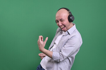 Asian bald man listening music with headphones while doing guitar gesture on green background