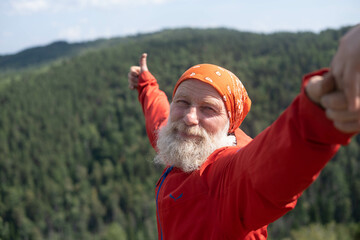a white elderly man takes a selfie on the top of a mountain, against the background of the sky and the forest from a bird's-eye view. The face of a mature gentleman with a red bandana on his head.