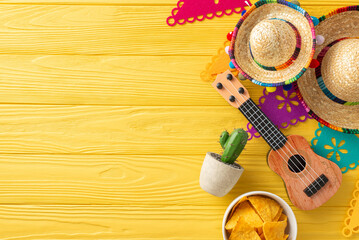 Cinco de Mayo top view scene: sombreros, vihuela, and a cactus on display. Colorful flag garland, and spicy nacho adorn the yellow wooden desk. Space for text