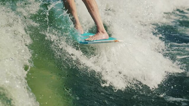 Wakesurfing. A man who is a beginner wakesurfer glides through the water behind a boat. Close-up of the board and legs.