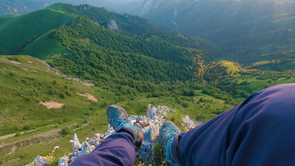 First-person view of legs of a man sitting on edge of a cliff overlooking a stunning mountainous landscape. The man is wearing blue pants and blue shoes, enjoying beauty of green hills and forests.