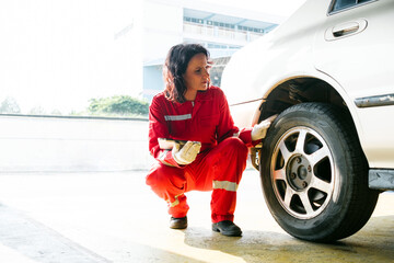 Portrait of a Caucasian female mechanic in a red uniform sitting and inspecting back wheel and tire on the right side of the car in the garage. Car repair service. Vehicle maintenance - 774658437