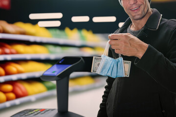 Closeup photo of male customer at supermarket self-checkout area holding money in medical mask. Automated checkout: Cash dollars exchanged at self-service terminal simplify supermarket purchases