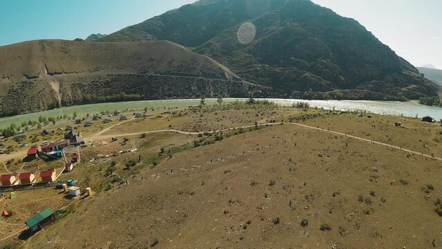 Majestic waters flow between undulating terrain. Serene presence of campsite along bank under benevolent gaze of clear sunny sky. River currents
