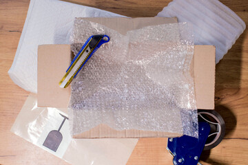 Open box with wrapped items, adhesive tape, scissors, paper and bubble wrap on wooden table, flat lay - obrazy, fototapety, plakaty