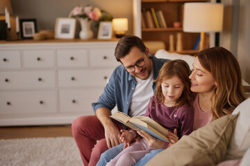Family with little daughter reading book on sofa in living room - obrazy, fototapety, plakaty