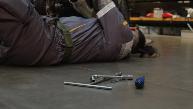 Multicultural mechanic in gray uniform changing instruments while lying on floor when repairing motorbike