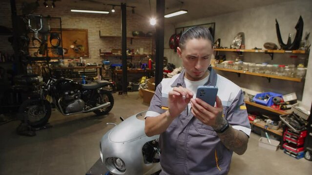 Medium shot of male mechanic ordering equipment for broken motorbike online via smartphone while working in garage