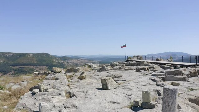 Approaching drone shot showing a flag being blown by the wind at the end of a viewpoint platform on the ancient historical site of of Perperikon, located in the province of Kardzhali, in Bulgaria.