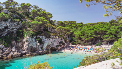 Turquoise shallow clear water, white fine sand beach and rocky cliffs with green pine trees, picturesque landscape of Cala Macarelleta in Menorca Island Spain, crowded with tourists in summer holiday