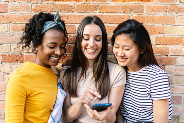 Happy young girls using mobile phone standing over brick wall background outdoors. Social media...