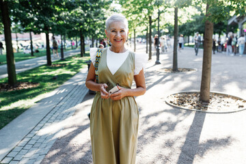 Pretty gorgeous fashionable mature female with short hair in stylish clothes standing with smartphone in hands, walking down city park after work day at office, looking at camera with smile - 774713036
