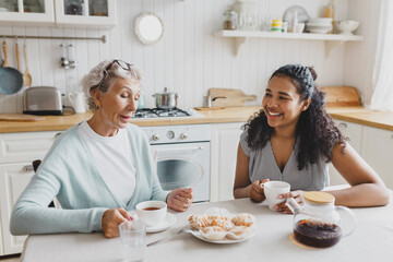 Indoor portrait of two people, black young female volunteer visiting with lonely retired female with gray hair, having nice conversation sitting at kitchen table, drinking tea with sweets - 774713234