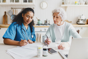 Elderly lady sitting at kitchen table near african american nurse in blue uniform writing prescriptions after giving her first aid, senior woman looking attentively at notes, holding bottle of water