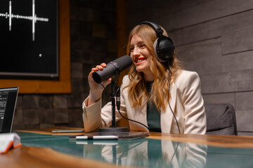 A woman with headphones is chatting into a microphone while sitting at a table. Podcast studio...