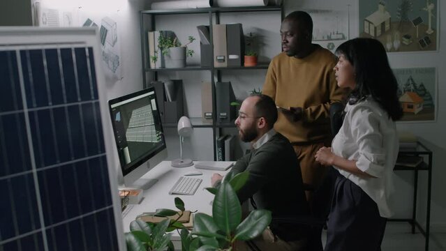Side pull out footage of group of diverse solar power engineers working on enhancing renewable energy technologies and discussing solar panel mock-up on computer screen in office