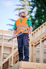 Carpenter building wooden frame two-story house. Bearded man in glasses holding a level, wearing protective helmet, overalls and orange vest. Concept of modern ecological construction.