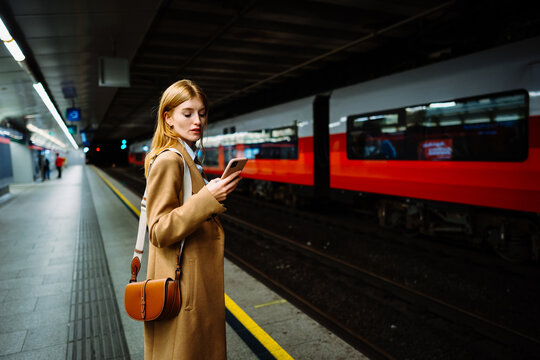Young serious businesswoman in suit surfing wireless internet on smartphone in metro